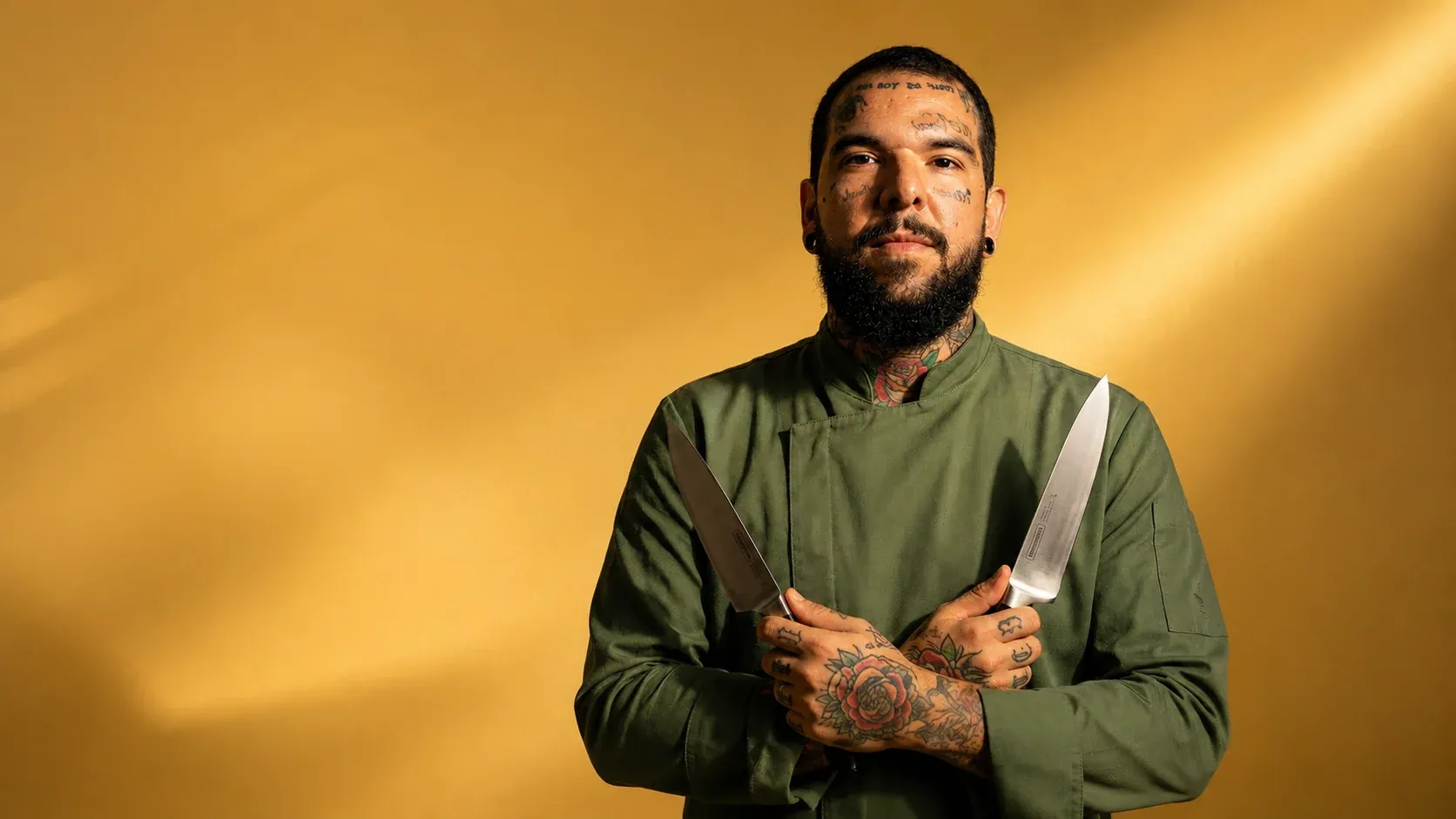 Fellipe Ferraz de dólmã verde-oliva, segurando duas facas de chef cruzadas diante de uma parede dourada